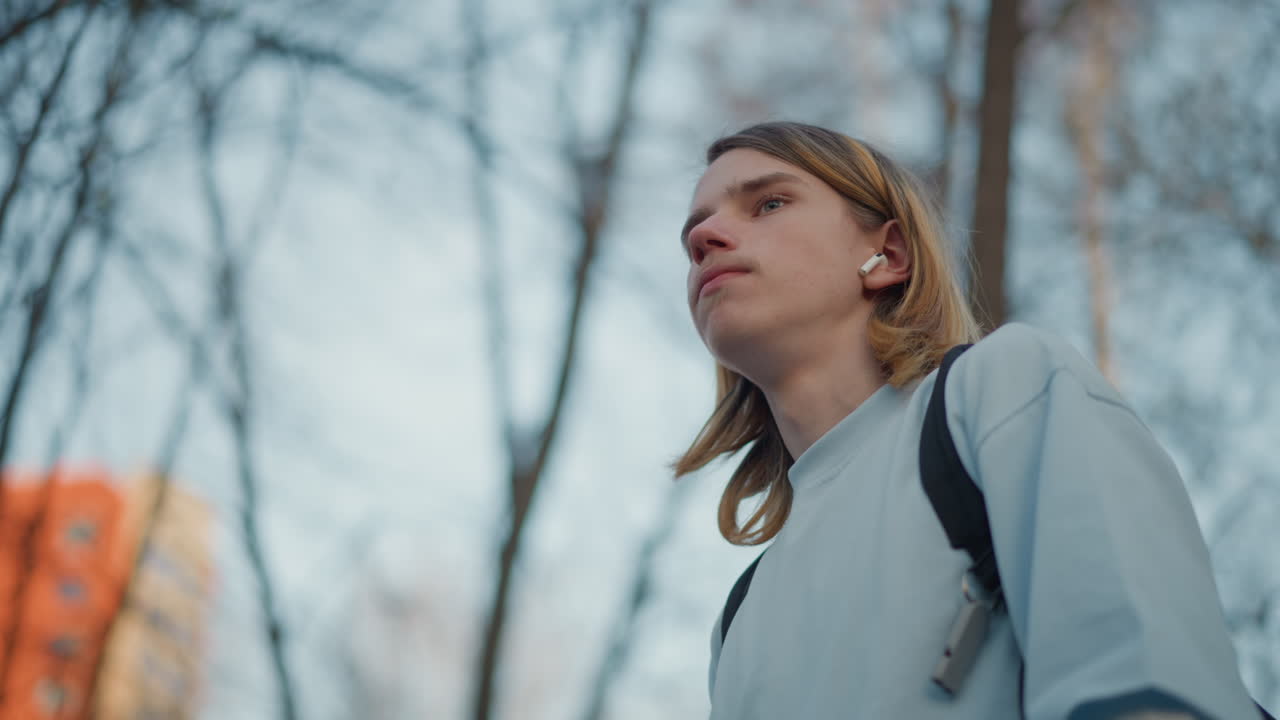 adolescente blanco bebiendo café para llevar con confianza bajo altos árboles en un parque urbano, toma en ángulo bajo que enfatiza la postura y los auriculares, el brillo cálido del atardecer y los edificios distantes realzan el ambiente relajado y audaz