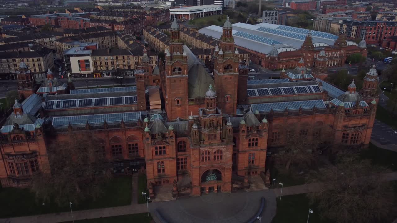 Drone captures Kelvingrove Art Gallery and Museum in Glasgow surrounded by urban rooftops and streets during peaceful evening light, showing stunning historic architecture and vibrant city layout