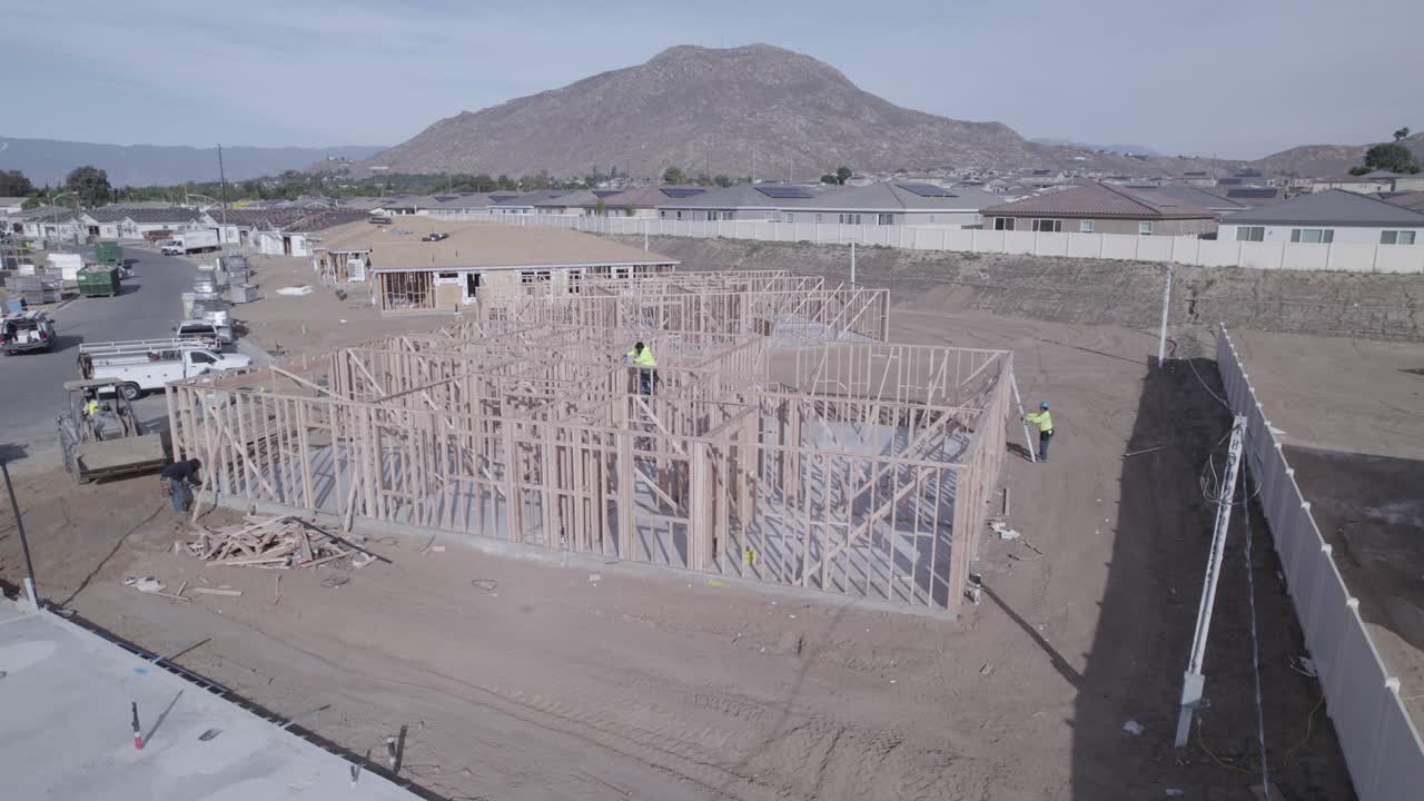 A drone progressively approaches a partially built house with only raised wall frames, highlighting a construction worker in the center diligently nailing all the frames together
