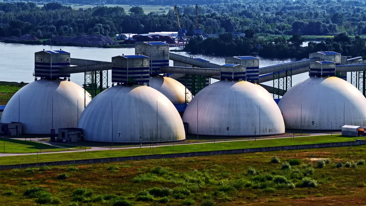 Una fila de grandes unidades de almacenamiento industrial con forma de cúpula se alza junto a una instalación ribereña, conectadas por sistemas de transporte elevados, con campos verdes y árboles distantes que forman un telón de fondo tranquilo.