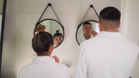 lovers brush teeth in front of hanging round mirrors in bathroom. couple in terry bathrobes conducts evening care routine standing together. hygienic routine