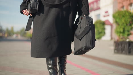 back view of person walking confidently on sunlit urban street, wearing chic black coat and leather boots, carrying two bags, blurred background shows building and decorative plant