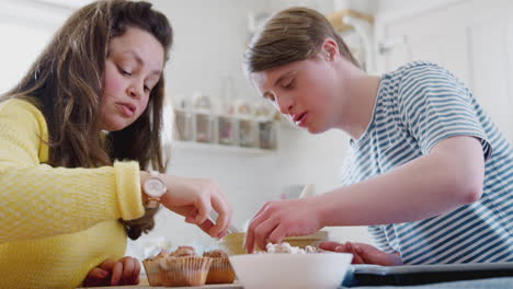 young downs syndrome couple decorating homemade cupcakes with marshmallows in kitchen at home