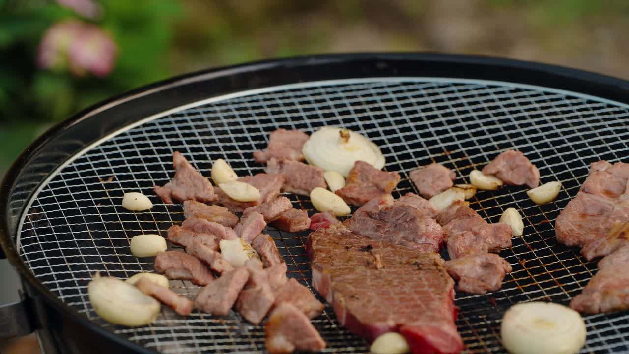 A close-up shot of tongs flipping a fresh, raw marbled beef steak for the first time on a hot charcoal grill during a Korean-style outdoor garden barbecue party