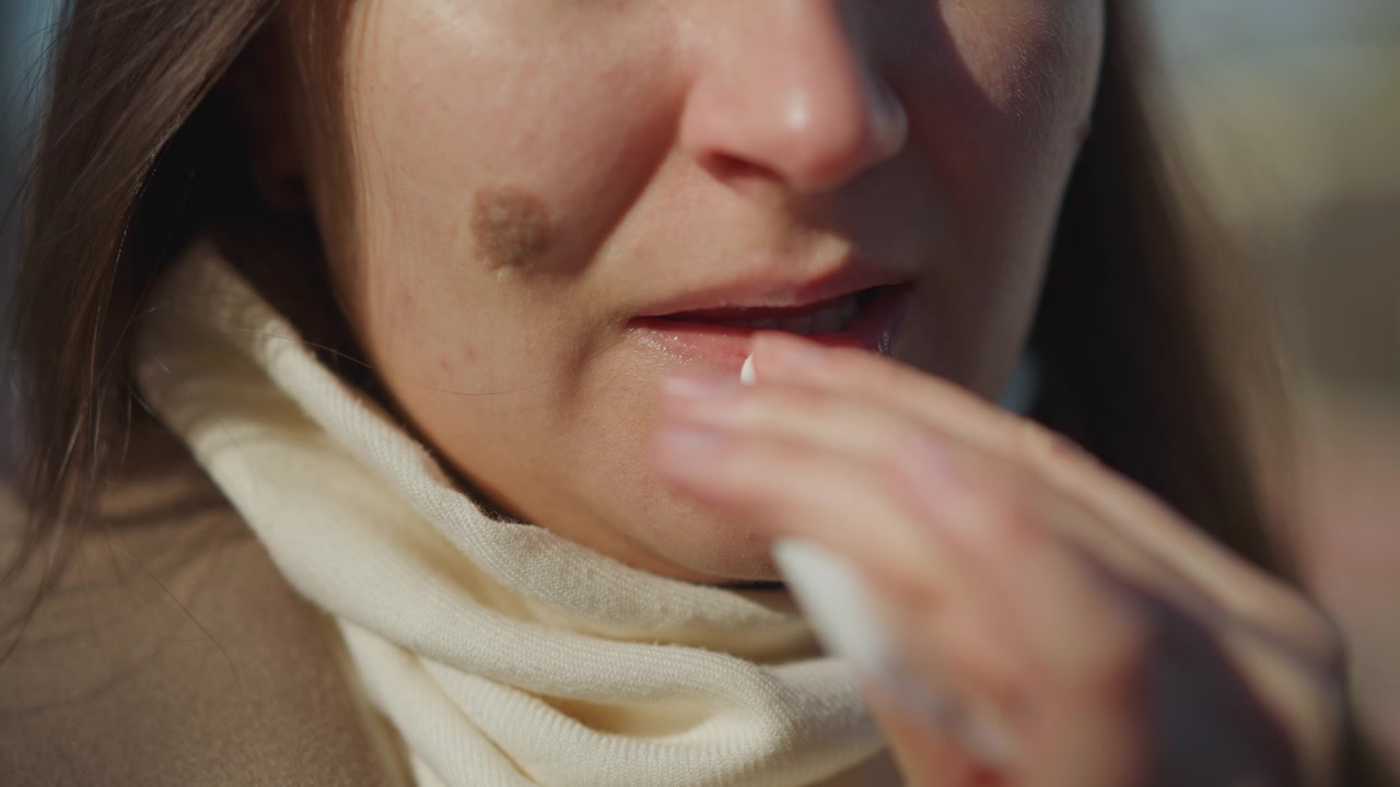 Primo piano di una donna che applica balsamo labbra sulle labbra mentre indossa una sciarpa color crema e un top marrone. L'azione è eseguita con movimenti costanti e un'illuminazione soffusa che si concentra sui dettagli della texture della pelle e del tessuto.