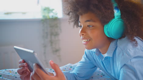 boy in bedroom lying on bed wearing headphones using digital tablet