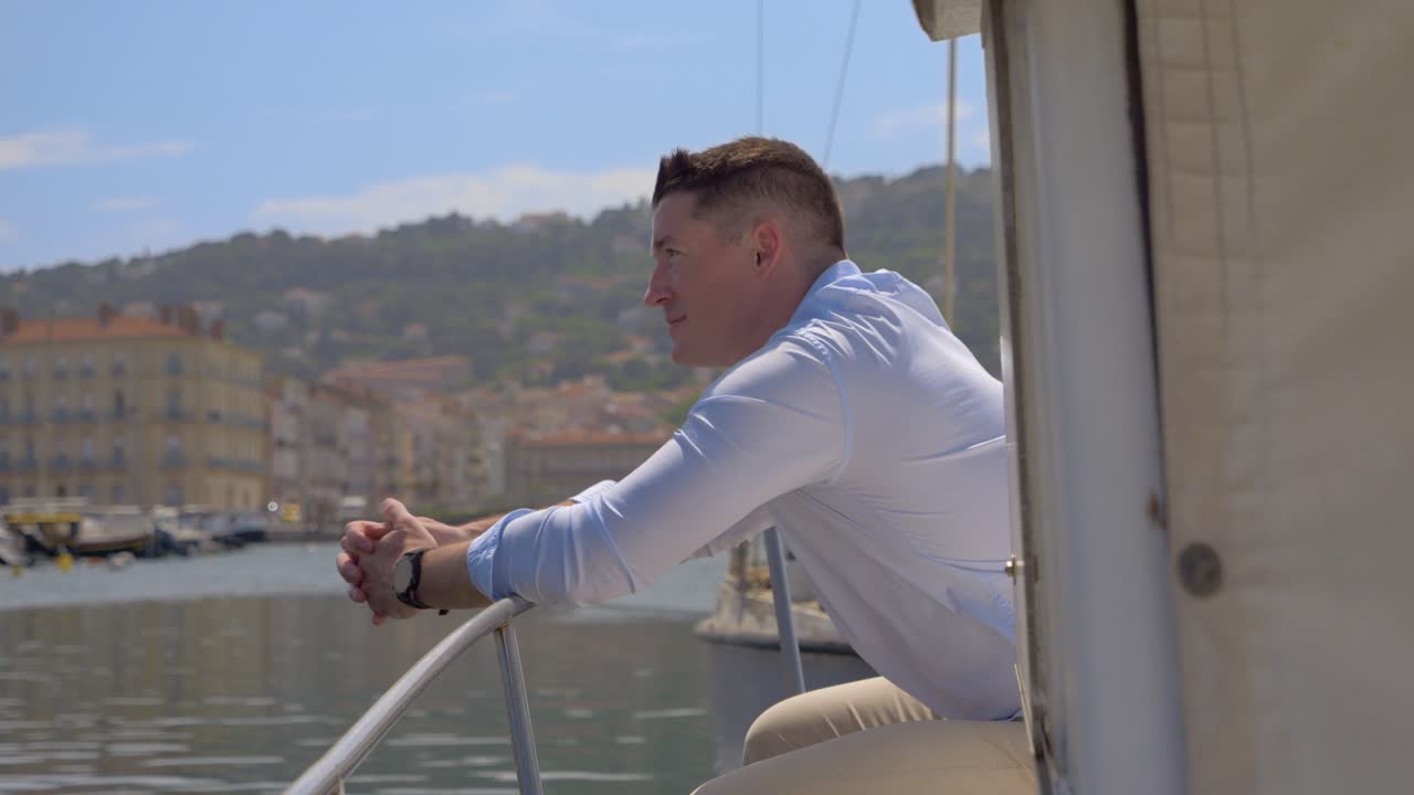 Un giovane uomo caucasico si appoggia al bordo della sua piccola barca e guarda il porto con un sorriso.