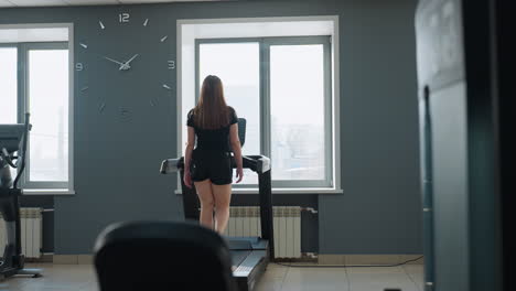 young woman in sportswear working out on treadmill facing window with sunlight streaming in, modern gym interior with second treadmill display blinking in background