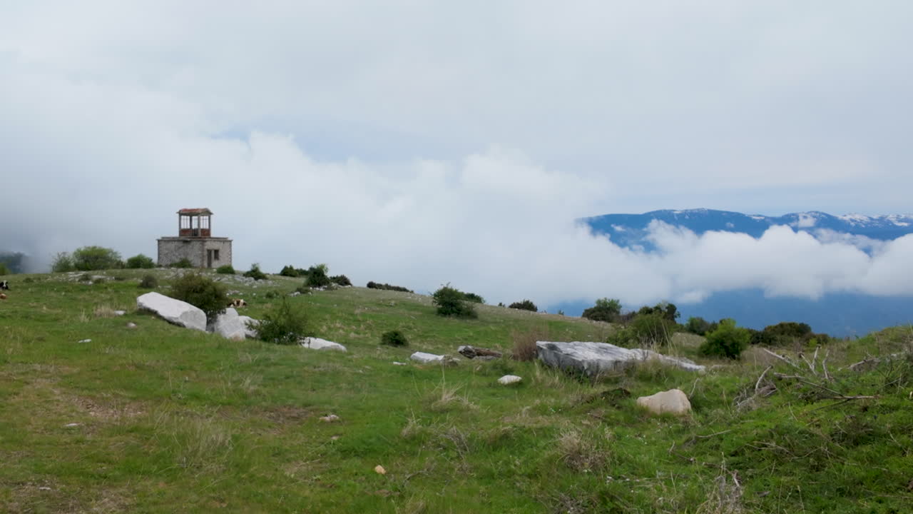 Un’inquadratura cinematografica di una solitaria struttura in pietra in cima a una montagna nebbiosa in Grecia, circondata da nuvole basse e prati alpini verdi