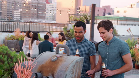 two men cooking barbecue for friends gathered on rooftop terrace with city skyline in background