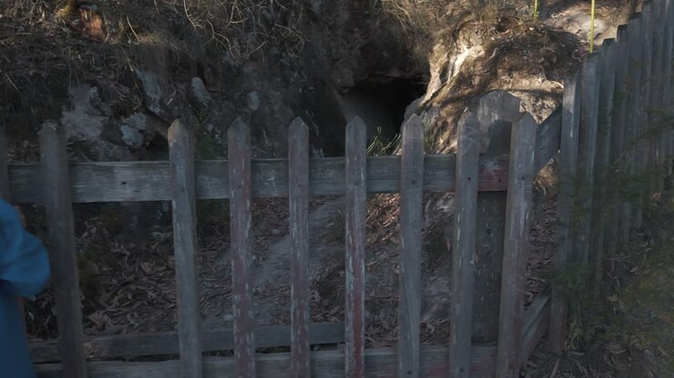 A person stands by a fenced entrance to an old mining cave at Warrandyte, Melbourne. Sunlight casts shadows on the rugged terrain, highlighting the cave's historical allure