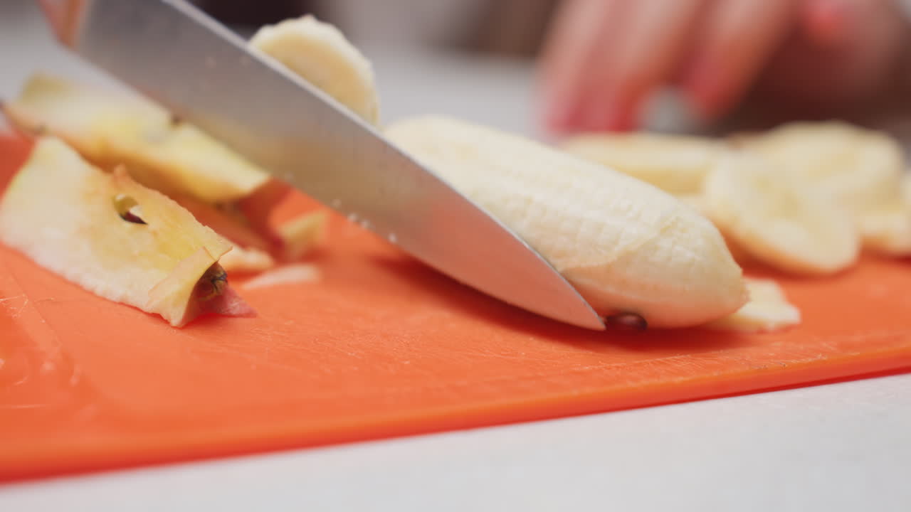 Close up female hand slicing banana with knife on orange rubber board, round pieces forming, texture visible, blurred background, kitchen prep, food hygiene focus, fresh fruit for snack