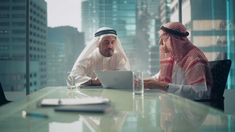dos empresarios emiratíes exitosos en kandura tradicional blanca sentados en una reunión de oficina, negociando y hablando sobre oportunidades de inversión, usando una computadora portátil.