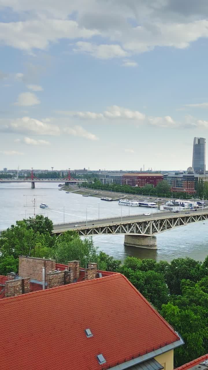 Vertical aerial drone shot over Budapest, featuring the Danube River, a busy bridge with moving cars, and a boat passing underneath.