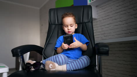 charming caucasian child in blue t-shirt sits in leather chair. baby boy plays with a smartphone.