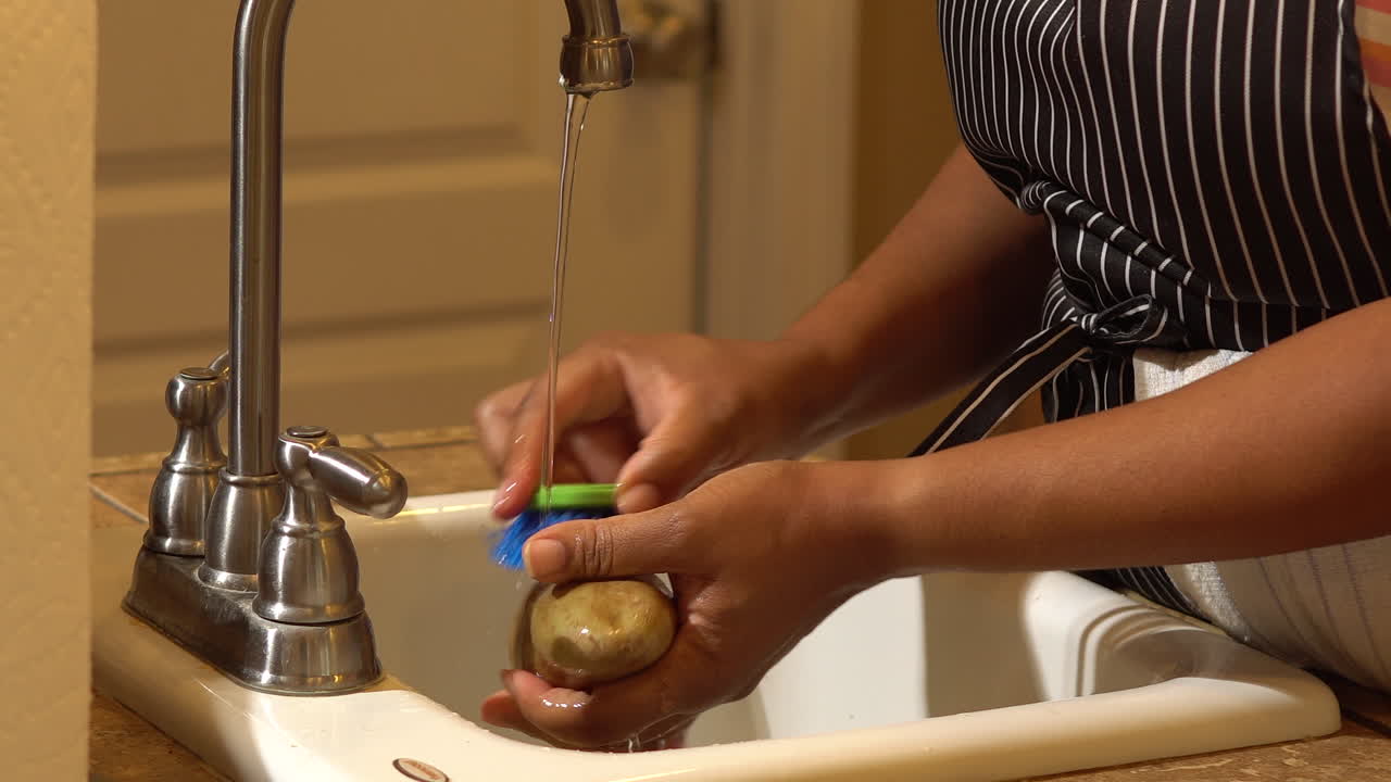 Black woman scrubbing potato with brush under water from kitchen sink, CLOSEUP