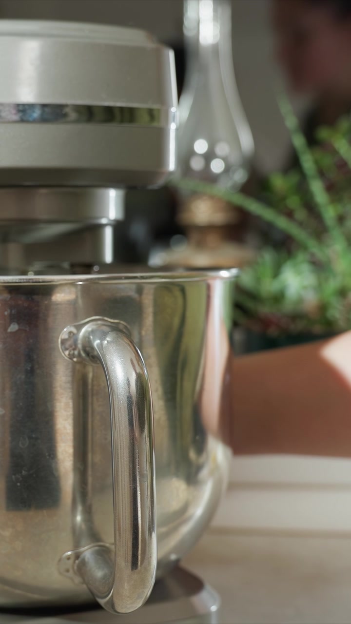 Close up of metallic kitchen mixer handle. Hand intermittently appears in background reaching towards or adjusting out of focus green plant. Blurred glass bottle and plants are