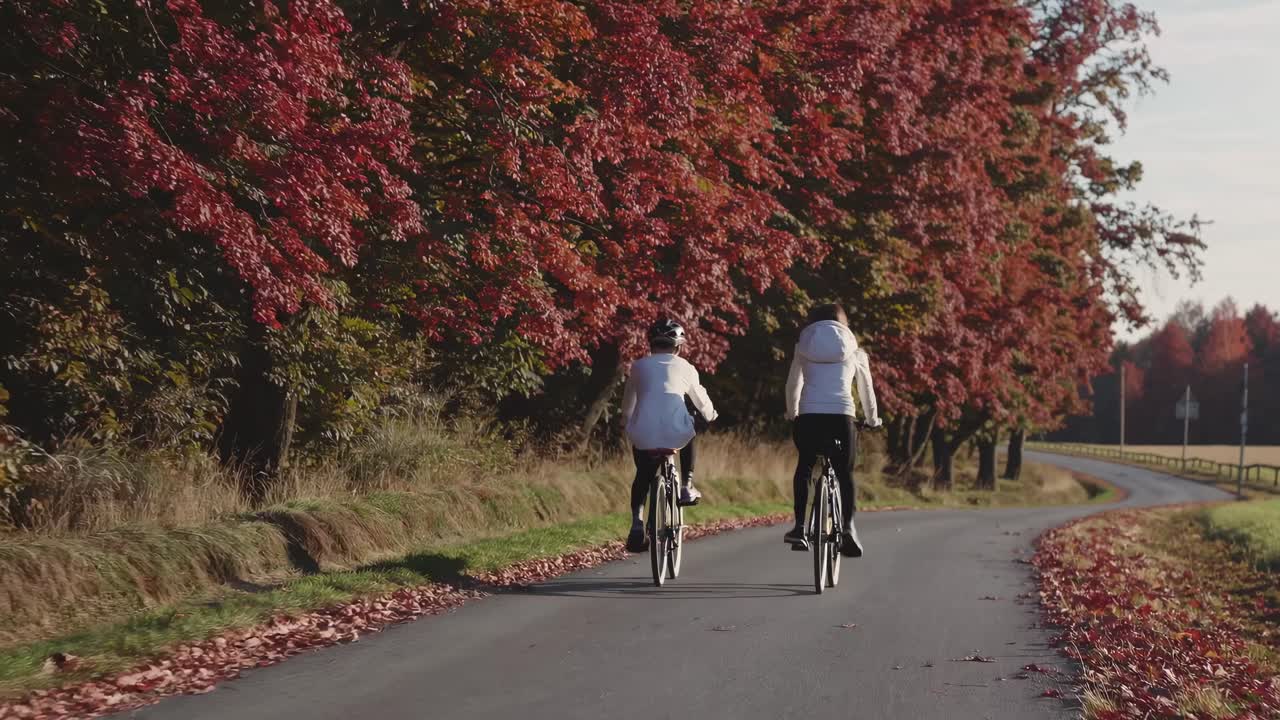 A scenic video captures two cyclists from behind, riding along a tree-lined road in autumn