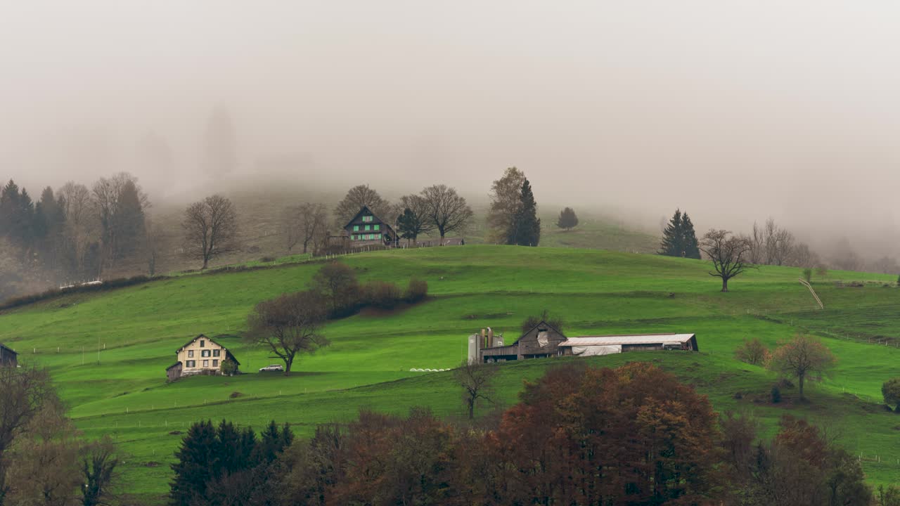 This still timelapse captures a misty morning in Schänis, St. Gallen, Switzerland. A traditional Swiss farmhouse sits atop rolling green hills, partially veiled by drifting fog.