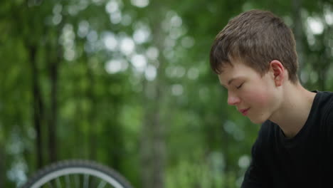 boy in black top thoughtfully lowers his head while sitting outdoors, with a blurred view of bicycle tire beside him, the serene background features blurred trees