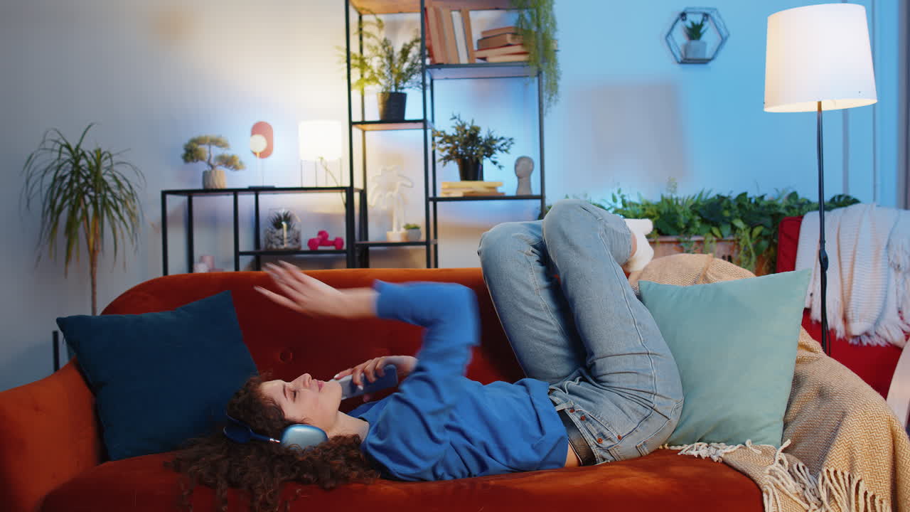 Joyful relaxed woman listening to smartphone music through wireless headphones lying on sofa dancing