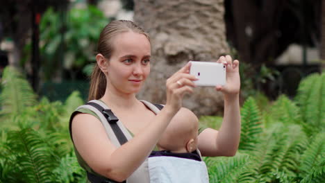 While-strolling-with-her-baby-in-a-kangaroo-backpack,-a-young-woman-documents-her-travels-using-a-mobile-phone.-She-captures-moments-and-looks-at-the-screen