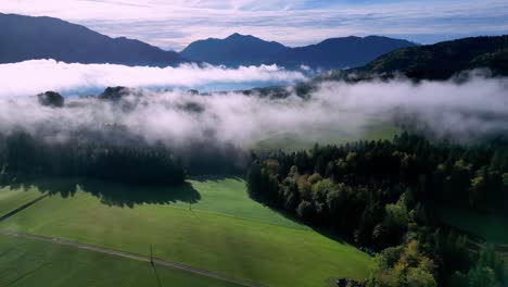 aerial view of a lush green valley filled with fog, with a winding lake snaking through the center and snow-capped mountains rising in the distance