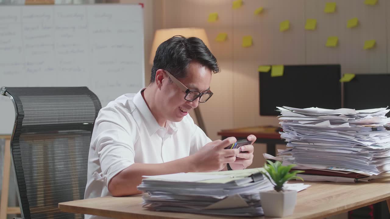 Happy Asian Man Celebrating Winning Game On Smartphone After Working With Documents At The Office