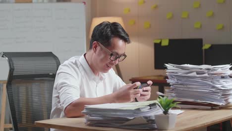 happy asian man celebrating winning game on smartphone after working with documents at the office