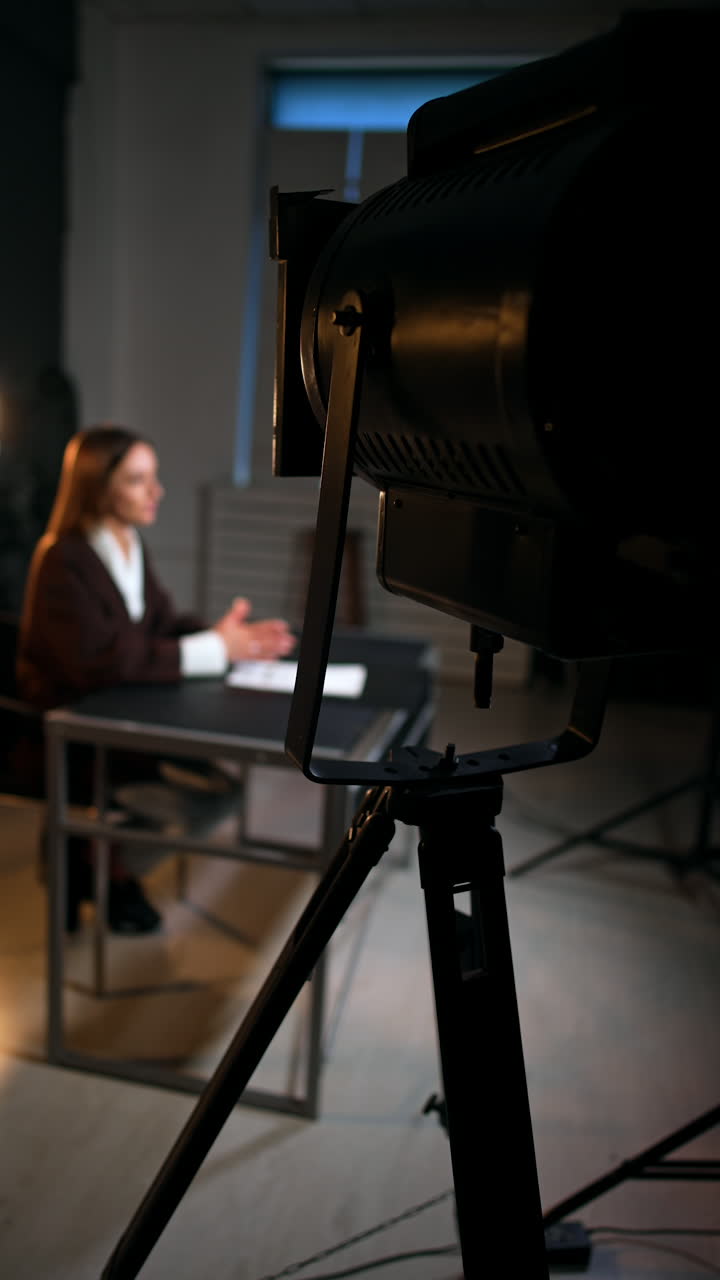 Professional soffit standing in the photo studio directed at the woman sitting at desk. Female reporter recording a video for blog. Vertical video.