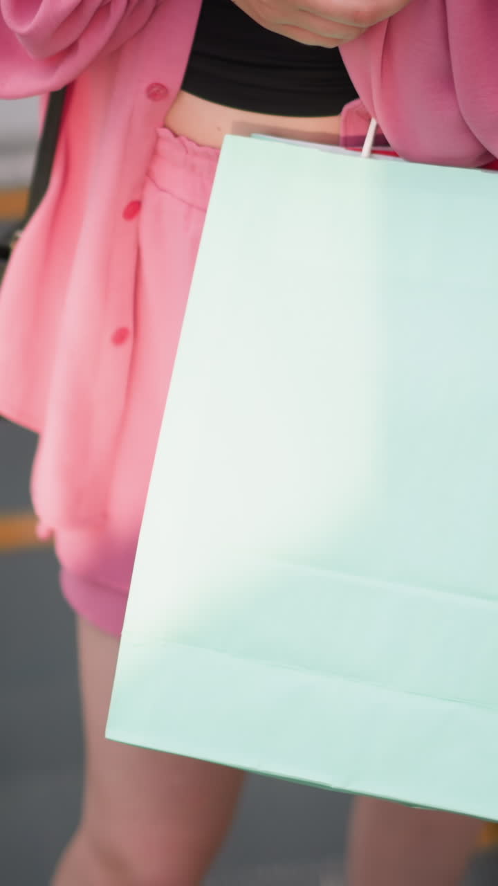 dama vestida con un vestido rosa sosteniendo bolsas de compras verdes y rojas en una escalera mecánica en movimiento, rodeada de bordes de vidrio, se para casualmente, llevando sus bolsas mientras la escalera se mueve