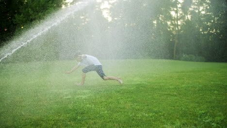 Happy-boy-playing-with-water-sprinkler-in-field.-Positive-guy-jumping-in-air