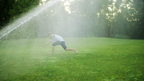 happy boy playing with water sprinkler in field. positive guy jumping in air