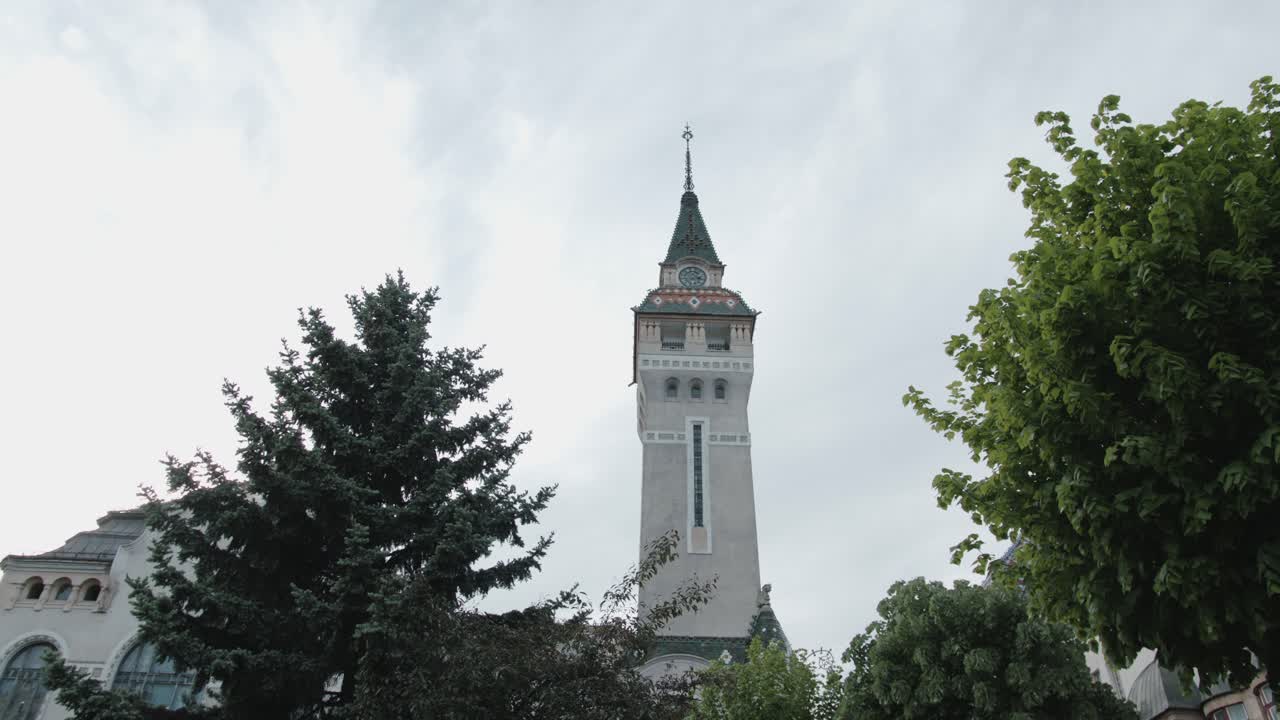 A static, low-angle shot of a historic European clock tower (Palace of Culture, Targu Mures) framed by trees. The ornate spire stands tall against a dramatic, grey, overcast sky