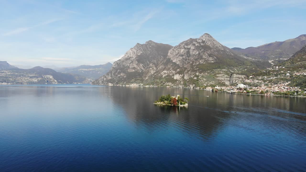 Drone ruota sopra il lago d'Iseo mostrando lo splendido panorama con una barca e una piccola isola durante una soleggiata giornata autunnale.