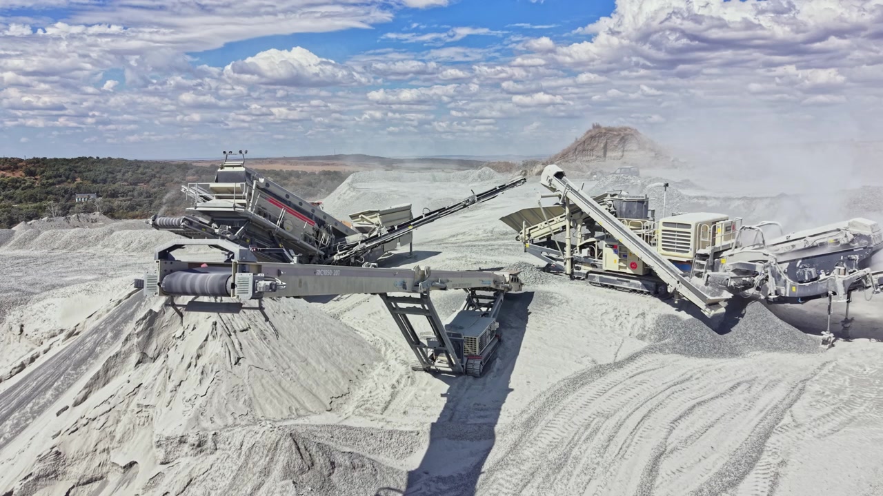Heavy machinery operates in a quarry with piles of material and blue skies in the background