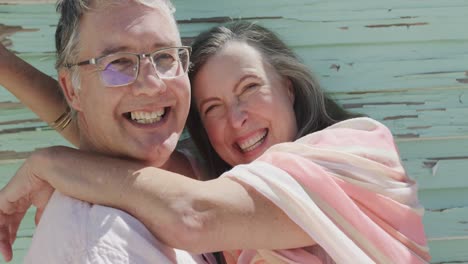 Portrait-of-happy-senior-caucasian-couple-embracing-and-laughing-by-wooden-wall,-in-slow-motion