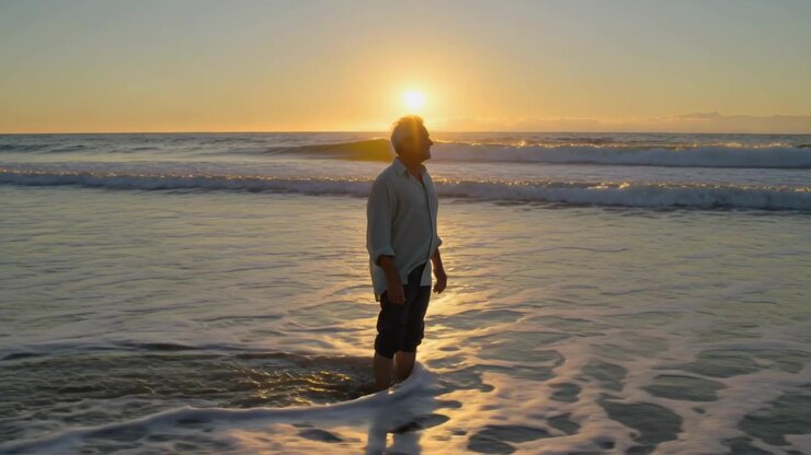 Sol se aproximando do horizonte, homem de camisa clara e shorts escuros caminhando em águas rasas, de frente para o sol.