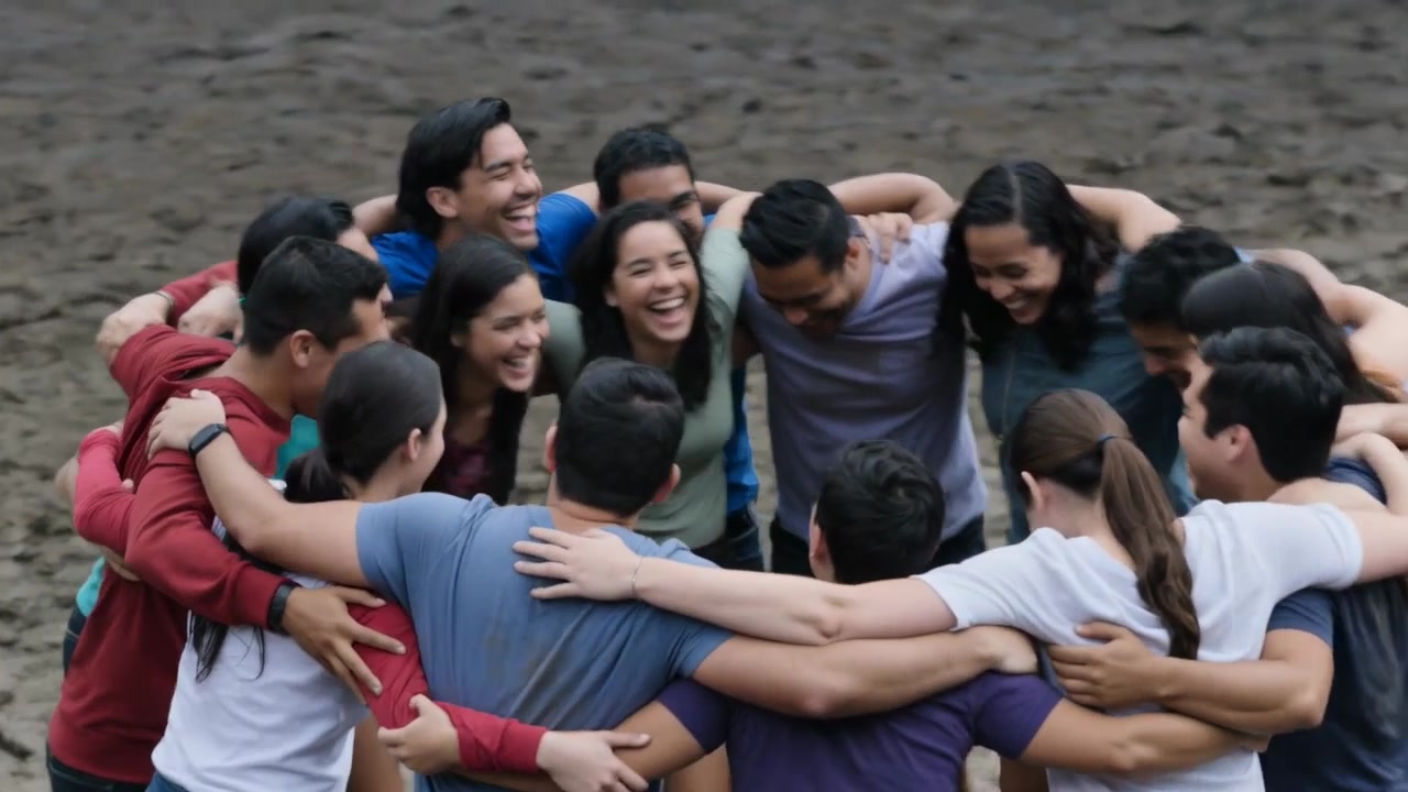 Laughing team linking arms, tightening huddle on muddy field for unity, casual tees ponytail