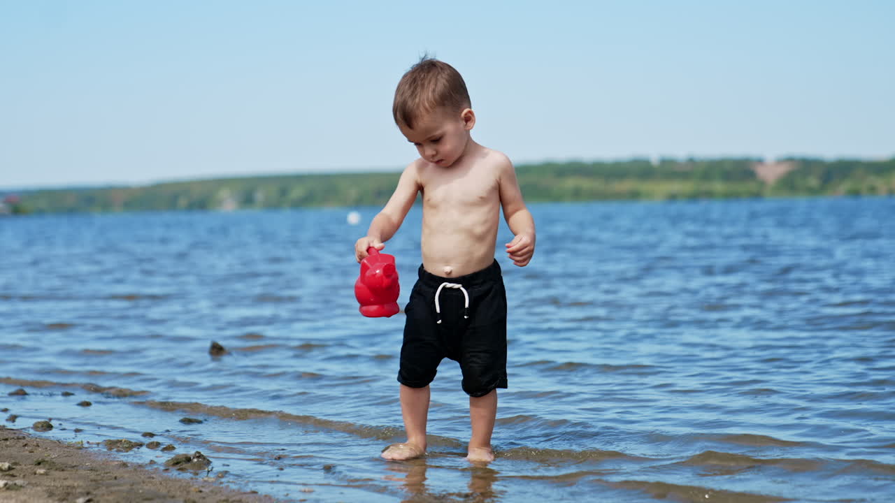 Beautiful Caucasian toddler playing in the beach. Kid carries a watering can filled with water from river.