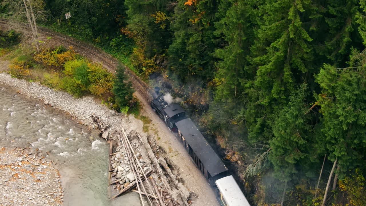 Aerial drone view of the moving steam train Mocanita in a valley along a river, hills covered with yellowing forest, Romania