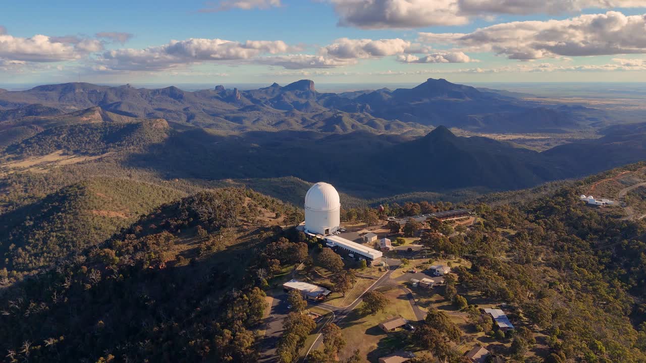 Aerial footage circles a large observatory dome atop a forested mountain ridge at sunset, revealing expansive views of rugged terrain and distant peaks