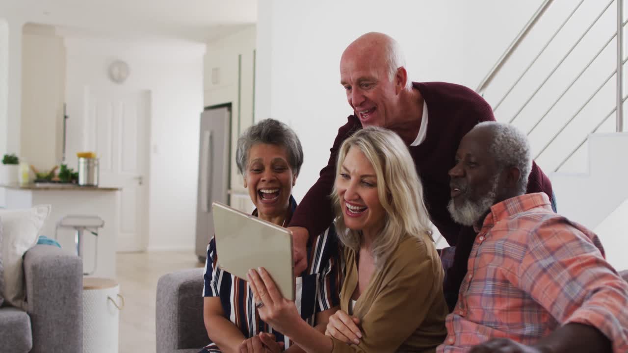 Two diverse senior couples sitting on a couch using a digital tablet and laughing