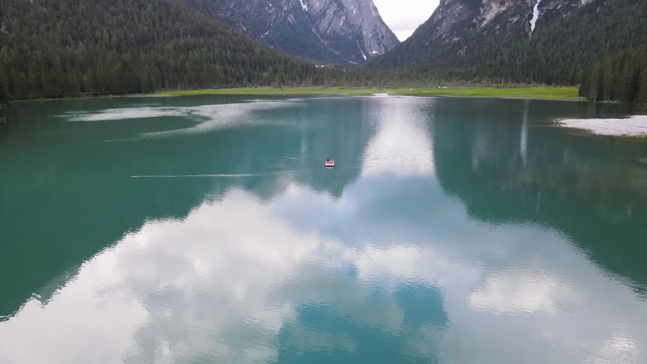 drone volando de un turista en un bote pequeño en medio del hermoso lago glacial reflectante rodeado de árboles en los dolomitas en italia