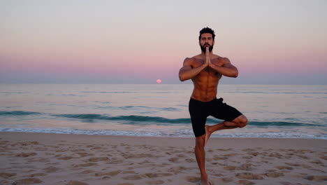 man practices yoga in tree pose on a beach at sunset