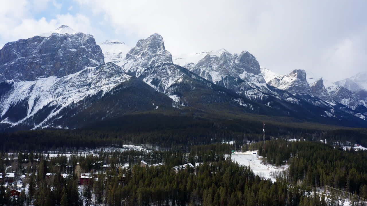 Aerial leftward glide reveals snow-draped Banff mountains and forest-lined cabins nestled deep in Canmore, Alberta’s winter wilderness.