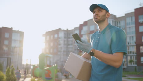 Un-Mensajero-Masculino-Está-Buscando-Un-Teléfono-Móvil-Para-Entregar-La-Dirección-De-Comida-De-Una-Tienda.-El-Repartidor-Navega-Por-Teléfono-En-La-Ciudad-Buscando-Direcciones-De-Entrega-De-Tiendas.-Entrega-Rápida