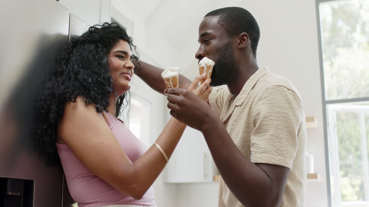 Coppia multietnica che si scambia coni gelato in cucina; l'uomo offre un cono, una sbavatura genera risate.