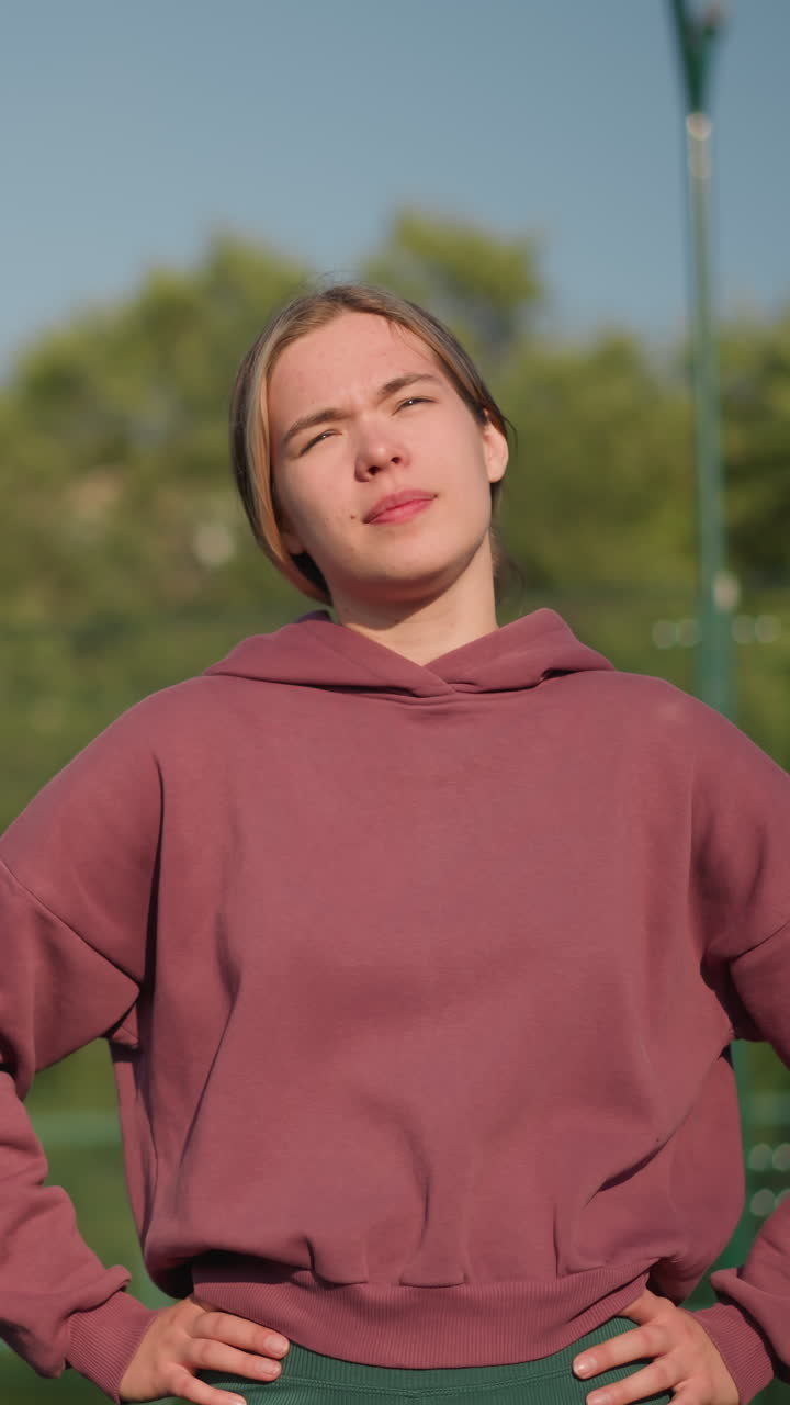 왼쪽에서 오른쪽으로 머리를 돌리는 동안 몸을 고있는 갈색 후디를 입은 여성, 태양빛이 그녀의 배경에 농구 허프와 함께 빛납니다.