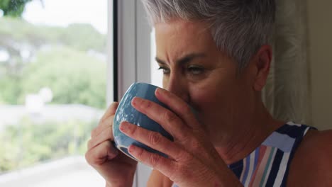 Caucasian-senior-woman-drinking-coffee-while-looking-out-of-the-window-at-home